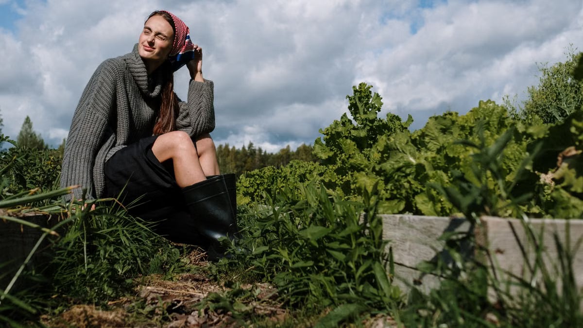 A caucasian woman wearing a gray sweater and bandana sits in a rural garden.